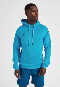 Blauwe vocht afvoerende hoodie met trekkoord in de capuchon, lange mouwen en logo op de borst. Gecombineerd met bijpassende korte broek. Gladde textuur.