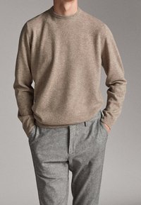 Beżowy sweter z długim rękawem zestawiony z jasnoszarymi spodniami, ręce swobodnie w kieszeniach, neutralne tło.