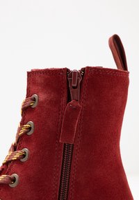 Botín de ante rojo hasta el tobillo con cremallera lateral, ojales de metal redondos y cordones amarillos y rojos en contraste. Textura suave con un ligero brillo.