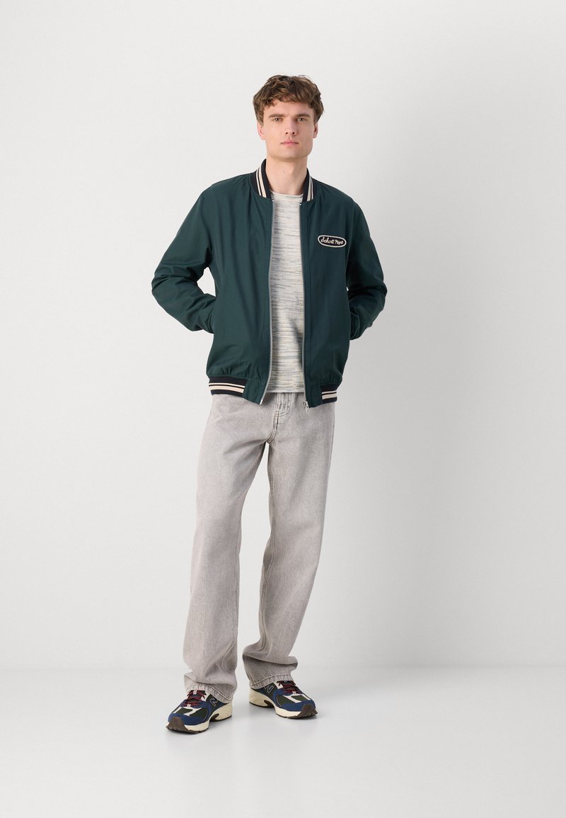 Veste bomber verte avec des poignets et un col côtelés rayés, portée sur une chemise grise à rayures, associée à un jeans gris clair et des baskets à motifs.