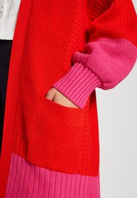 Ręka w kieszeni długiego, dzianego kardiganu w czerwono-różowych blokach kolorystycznych z prążkowanymi mankietami, noszonego na białym topie i ciemnych spodniach.