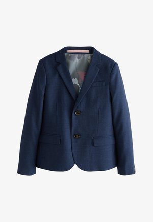 Junger blonder Junge trägt einen marineblauen Anzug, weißes Hemd und hellgraue Krawatte, steht und schaut leicht zur Seite.