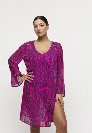 Vrouw met donker haar in een roze-zwart zebrapatroon jurk met lange mouwen en een hoge split aan de dij, staand met één hand op de heup.