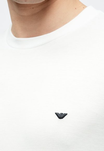 Fehér póló kerek nyakkivágással és rövid ujjakkal. Kis fekete hímzett logó található a bal mellrészén. Puha tapintású anyag.