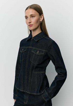 Γυναίκα με ίσια μαλλιά που φοράει σκούρο denim μπουφάν με κουμπωτές τσέπες στο στήθος και ταιριαστό denim παντελόνι, στέκεται μπροστά σε απλό φόντο.