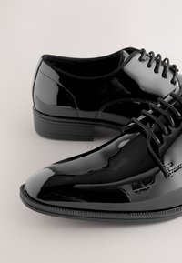 Paire de chaussures habillées en cuir verni noir brillant avec lacets, présentée sur un fond clair uni.
