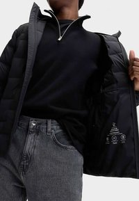 Veste noire matelassée avec fermeture éclair argentée, portée ouverte sur un haut noir. L'étiquette intérieure affiche les instructions d'entretien et les détails du matériau, en texte blanc.