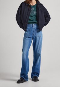 Dark, relaxed-fit jacket over a green sweatshirt. Wide-leg blue jeans with front pockets. Black glossy shoes. Smooth, casual textures.