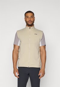 Gilet sans manches beige avec un col montant, fermeture éclair et accent logo noir. Chemise à manches courtes grise en dessous. Texture de tissu lisse.