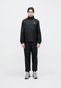 Fiatal férfi fekete Nike széldzsekit és hozzáillő nadrágot visel, szürke sportcipőben, fehér háttér előtt áll.