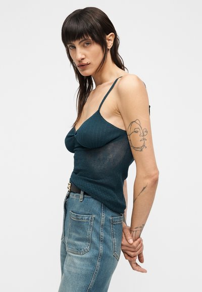 Πράσινη ριγωτή αμάνικη μπλούζα με λεπτές τιράντες, συνδυασμένη με ανοιχτόχρωμο τζιν παντελόνι με ξεθωριασμένη υφή. Ορατά εντυπωσιακά τατουάζ στα χέρια.