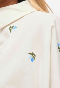 Gros plan sur un tissu crème avec un motif de petites baies bleues brodées et des feuilles vertes.