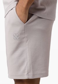 Ανοιχτό μπεζ σορτς με πλαϊνή τσέπη και διακριτικό κεντημένο λογότυπο στον μηρό, φορεμένο με το χέρι εν μέρει μέσα στην τσέπη.