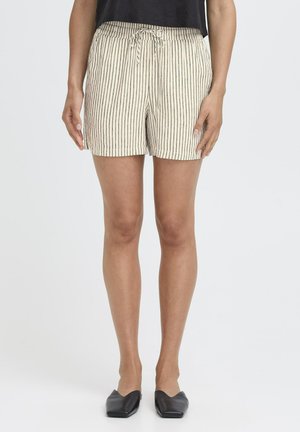 Femeie purtând pantaloni scurți bej cu dungi verticale negre și șnur, precum și pantofi negri comozi cu vârf ascuțit, stând pe fundal alb.