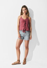 Top sin mangas de color rosa frambuesa con un delantero con volantes, combinado con pantalones cortos de mezclilla claros y sandalias de tiras marrones. Tejido suave, ajuste relajado.