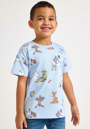 Smilende dreng iført lyseblå T-shirt med farverige tegneseriefigurer og naturmotiver, kombineret med blå jeans.