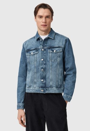 Jonge man met kort bruin haar, gekleed in een blauwe spijkerjack over een wit overhemd en zwarte broek, staand tegen een effen grijze achtergrond.