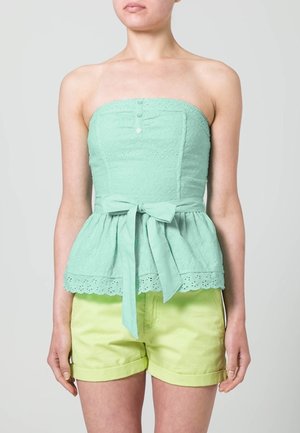 Top sans manches vert menthe avec texture œillets, design peplum, coutures verticales et un nœud à la taille ; associé à un short jaune clair.