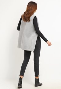 Szara kamizelka bez rękawów wykonana z miękkiego materiału, z długim tyłem i bocznymi rozcięciami. Nosi się ją na czarną bluzkę z długim rękawem, w zestawieniu z czarnymi dżinsami i butami.