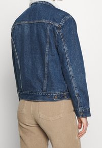 Jeansjacka i mörkblått med krage av syntetpäls. Har en klassisk design, knappstängning och synliga sömnadsdetaljer.