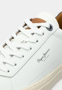 Baskets en cuir blanc avec lacets blancs, semelle beige, détail de languette couleur fauve et logo noir "Pepe Jeans London" sur le côté.