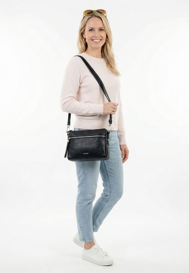 Lächelnde Frau mit hellrosa Pullover, blauen Jeans und weißen Turnschuhen, die eine schwarze Umhängetasche mit Reißverschluss trägt und goldene Creolenohrringe trägt.