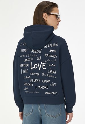 Person trägt einen dunkelblauen Hoodie mit dem Wort "love" in mehreren Sprachen auf dem Rücken, kombiniert mit blauen Jeans und Schildpatt-Brille.