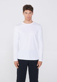 Langärmliges weißes Baumwoll-T-Shirt mit Rundhalsausschnitt, glatter Textur und tailliertem Design, kombiniert mit dunklen Hosen.