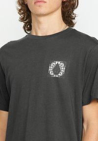 Sort bomulds t-shirt med et lille hvidt spindelvævsgrafik med en tåreformet figur i midten på venstre brystområde.