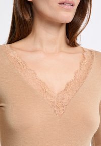 Top de manga larga beige con escote en V, que presenta un delicado borde de encaje alrededor del cuello. La tela tiene una textura suave y un diseño ajustado.