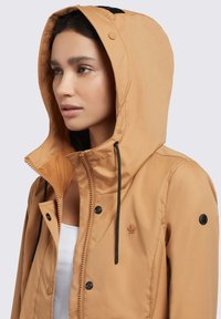 Svetlohnedá kapucňová bunda vyrobená z ľahkého materiálu. Obsahuje čierne šnúrky, zips na prednej strane a malé logo na hrudi.