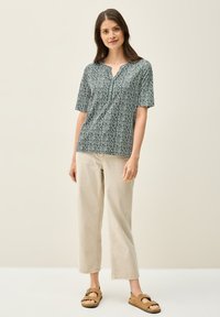 Kortärmad, grönmönstrad blus med V-ringning, kombinerad med beige, korta byxor och bruna sandaler. Slät tyg med geometrisk design.