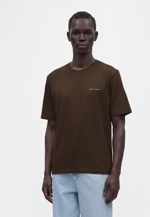 Mann trägt ein schlichtes braunes T-Shirt mit kurzen Ärmeln und kleinem "Tiger of Sweden"-Text, kombiniert mit hellblauen Jeans, steht vor weißem Hintergrund.