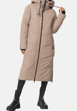 Frau trägt einen langen beige gepolsterten Mantel mit Kapuze, schwarzem Reißverschluss und schwarzen klobigen Stiefeln, hält eine Kordel in der Nähe ihres Gesichts.