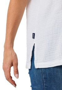 Weißes Baumwolloberteil mit strukturiertem, zerknittertem Design. Mit seitlichen Schlitzen und einem blauen Etikett mit der Aufschrift "SEASIDE". Kombiniert mit blauen Jeans.