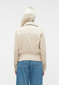 Osoba s dugom svijetlosmeškom kosom koja nosi bež jaknu i plave traperice, okrenuta leđima prema jednostavnoj bijeloj pozadini.