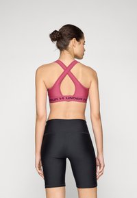 Roza sportska grudnjak s dizajnom leđa u obliku križa, s širokim crnim pojasom označenim "Under Armour". Crne kratke hlače s glatkim, prilagođenim izgledom.