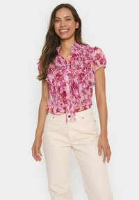 Short-sleeved blouse with a pink floral pattern, featuring ruffles down the front and a button closure. Fabric is sheer and lightweight.