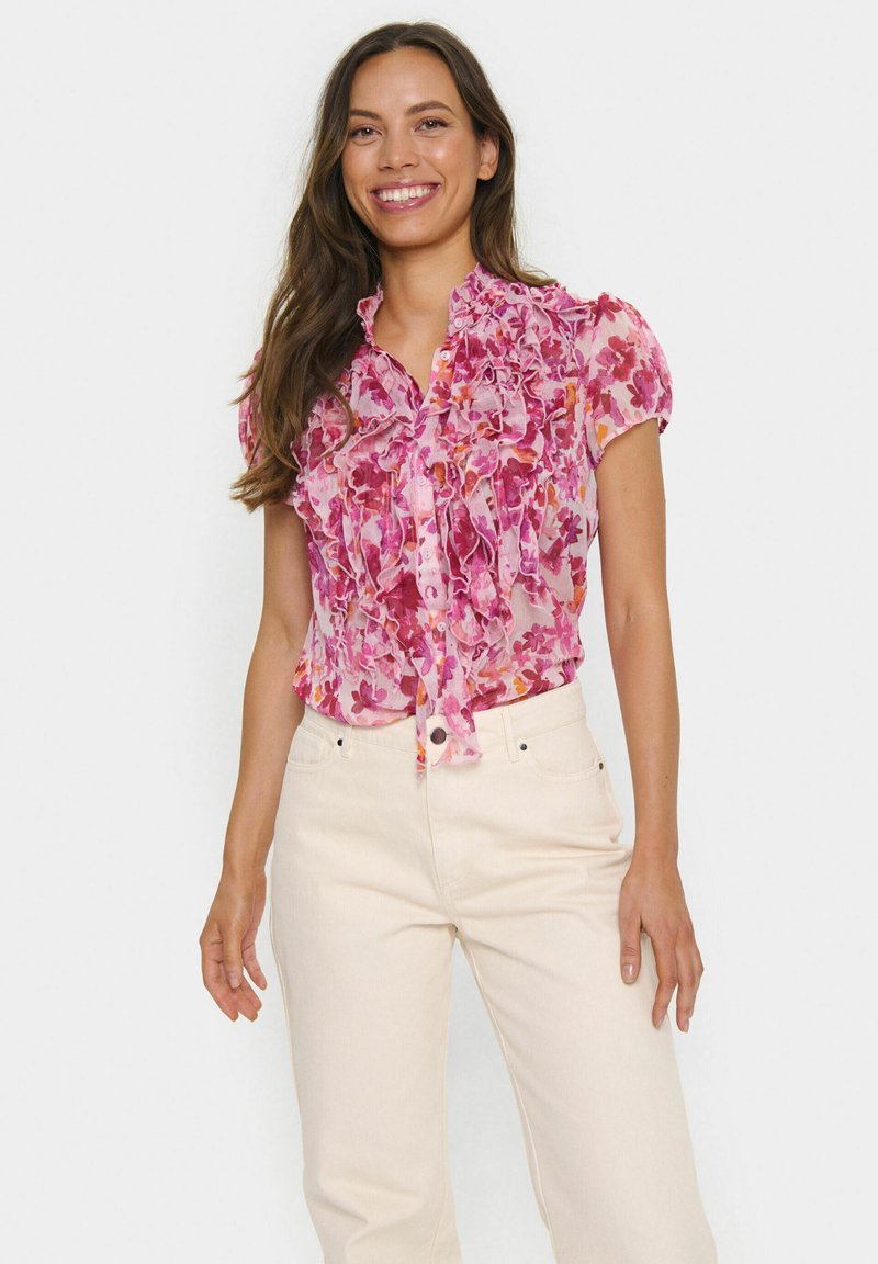 Short-sleeved blouse with a pink floral pattern, featuring ruffles down the front and a button closure. Fabric is sheer and lightweight.