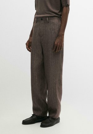 Mężczyzna ubrany w brązowe spodnie w paski o szerokich nogawkach, brązową koszulę z krótkim rękawem i czarne sznurowane buty, stojący na jednolitym tle.