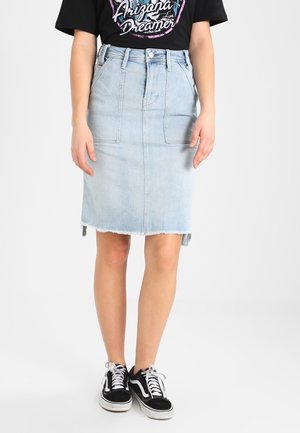 Femme portant une jupe en denim bleu clair longueur genou, un t-shirt graphique noir et des baskets décontractées noires et blanches, debout sur un fond blanc.