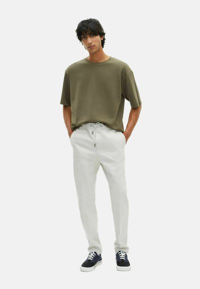 Giovane uomo con capelli scuri, che indossa una t-shirt oversize verde oliva, pantaloni bianchi con cordoncino e sneakers nere, in piedi con le mani in tasca.