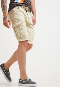 Shorts cargo beiges avec plusieurs poches, une ceinture et une coupe relaxée. Le tissu semble durable et a une texture lisse.