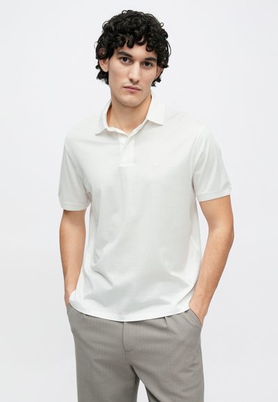 Bílá polokošile s límečkem, krátkými rukávy a jemným logem. Hladká textura, doplněná o světle šedé kalhoty.