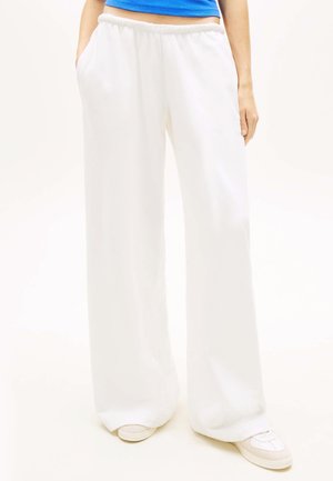 Person trägt weiße, weite Hosen, weiße Sneaker und ein blaues Crop-Top, steht mit den Händen in den Taschen.