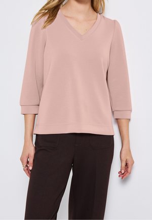 Vrouw die een lichtroze V-hals top met driekwartmouwen en donkerbruine broek draagt, tegen een effen lichte achtergrond.