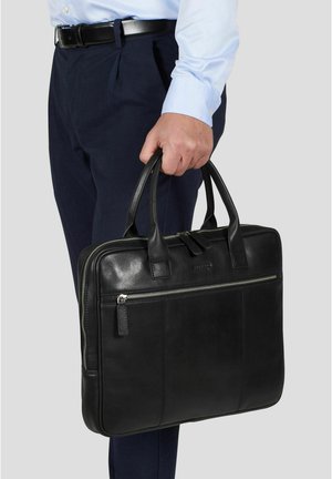 Mann i lys blå skjorte og mørke bukser holder svart skinnmappe med glidelåslomme mot grå bakgrunn.