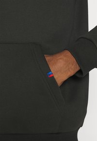 Main partiellement dans la poche avant d'un sweat-shirt vert foncé avec une petite étiquette en tissu rouge et bleue.