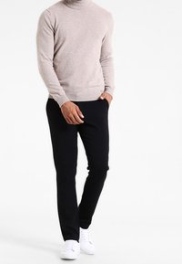 Ljusbeige polokragetröja i mjukt tyg, kombinerad med svarta slim fit-byxor och vita sneakers, på en enfärgad bakgrund.
