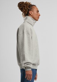 Sudadera gris claro con cuello alto, puños y dobladillo acanalados, hecha de tela suave, con un corte relajado y un diseño minimalista.